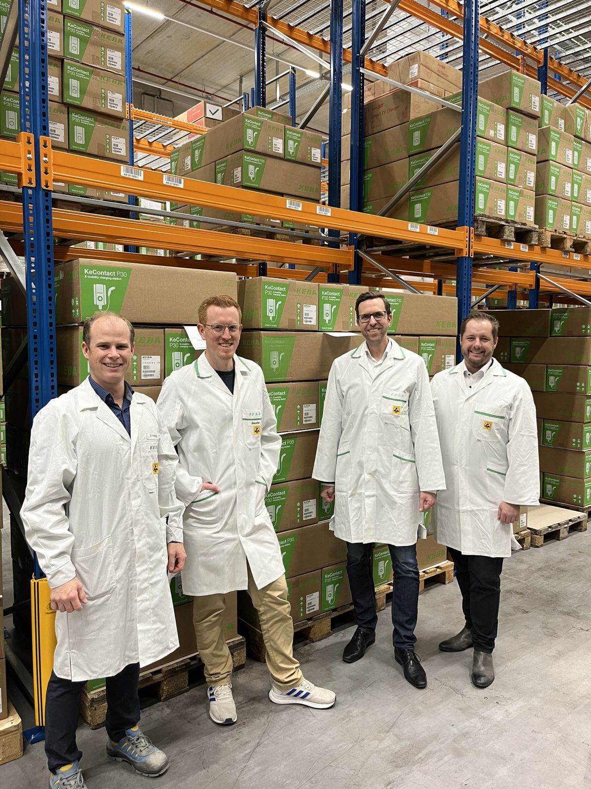 Four men in white lab coats stand in a warehouse surrounded by KEBA wallbox boxes.