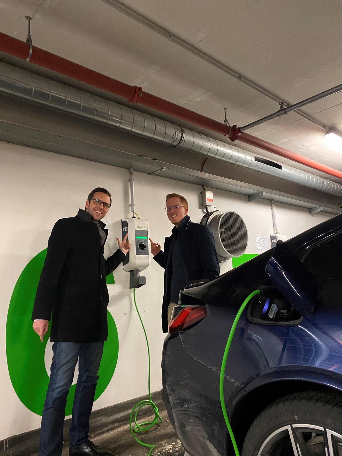 Two men stand next to an installed KEBA wallbox in an underground garage, pointing at the charging display.