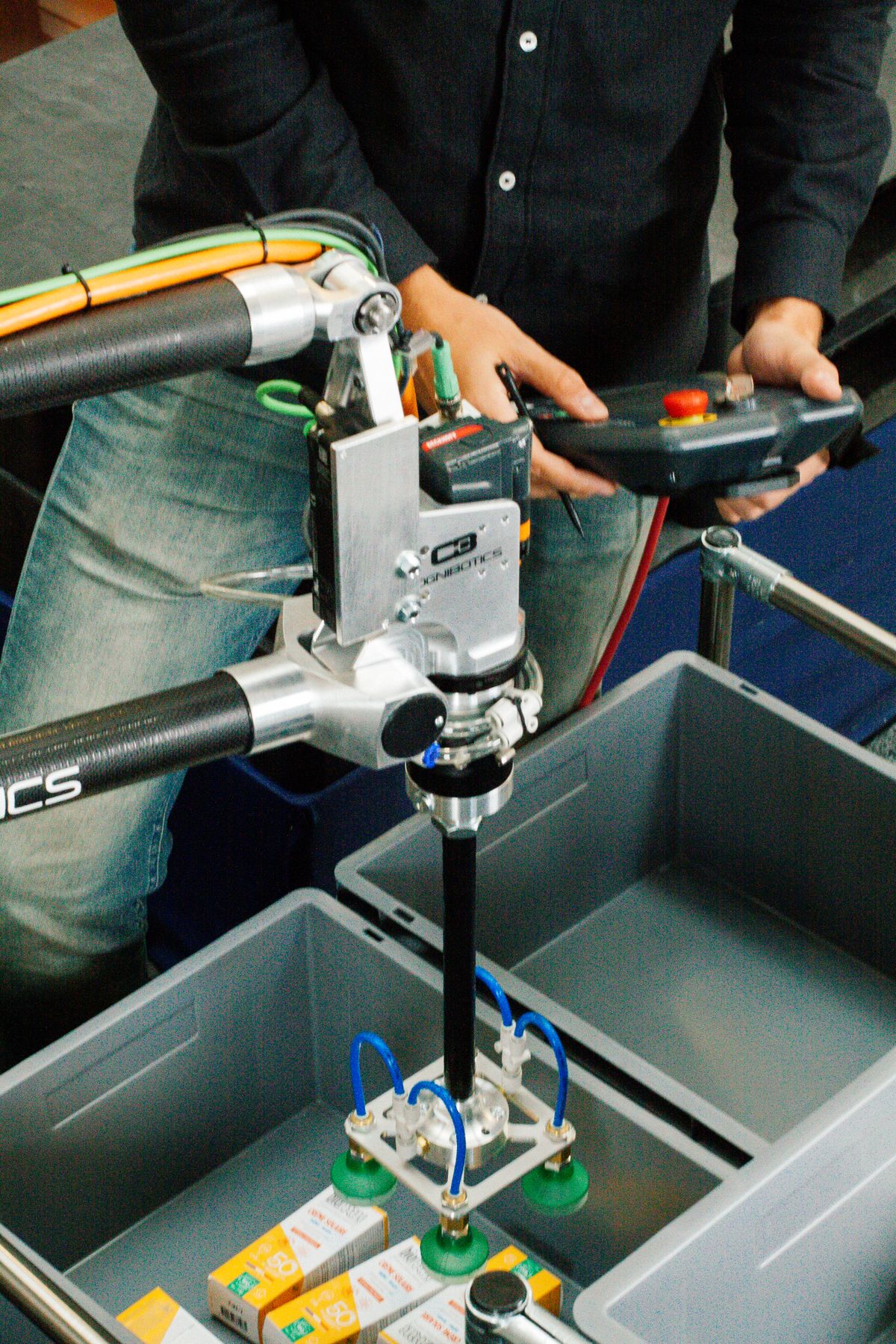 Ein Techniker steuert mit einem Handbediengerät einen Industrieroboter mit Vakuumgreifer, der Produktverpackungen präzise in Kunststoffboxen sortiert – Beispiel für automatisierte Materialhandhabung.