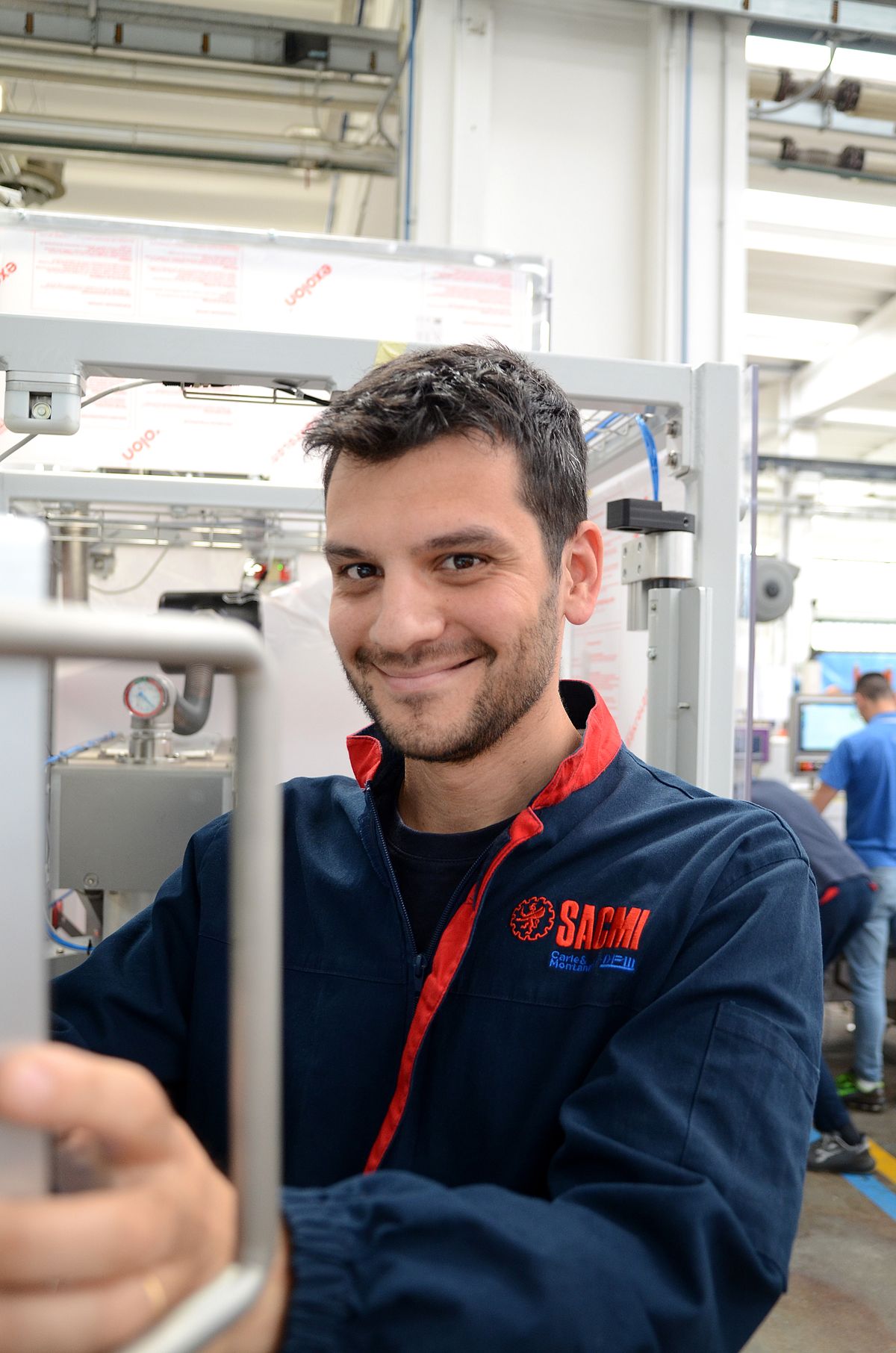 Ritratto di un tecnico in uno stabilimento produttivo che sorride alla fotocamera. Indossa abiti da lavoro con il logo rosso SACMI, simbolo di impegno e competenza nell’automazione industriale.