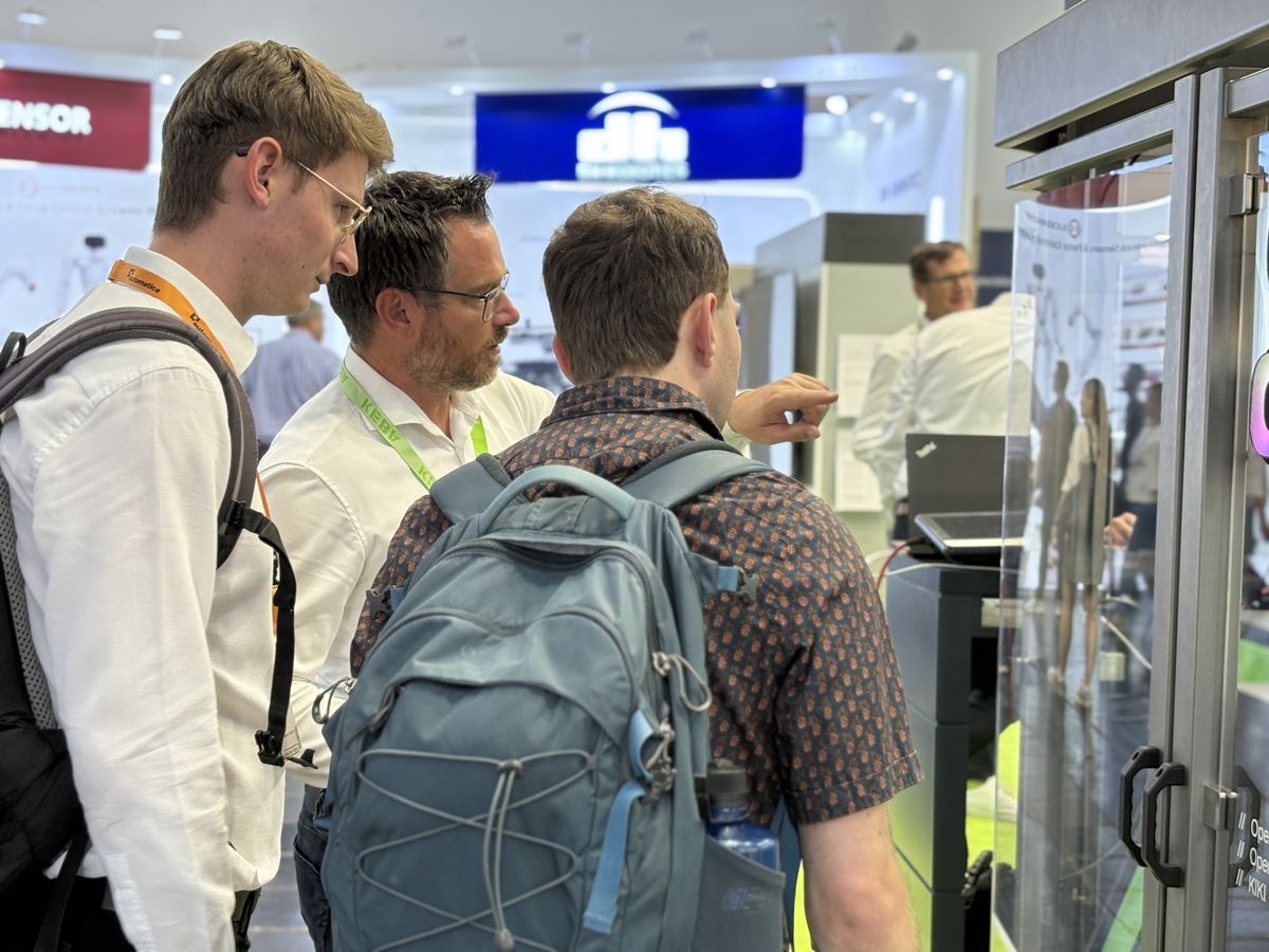 Drei Männer begutachten eine Roboterzelle am KEBA-Messestand. Einer erklärt technische Details und zeigt durch die Scheibe auf den Roboterarm.