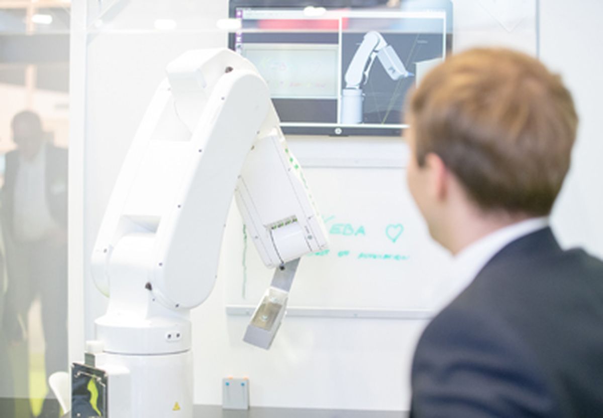 An industrial robot being demonstrated at a trade fair booth, while a person observes its motion and control via a graphical visualization on a screen.