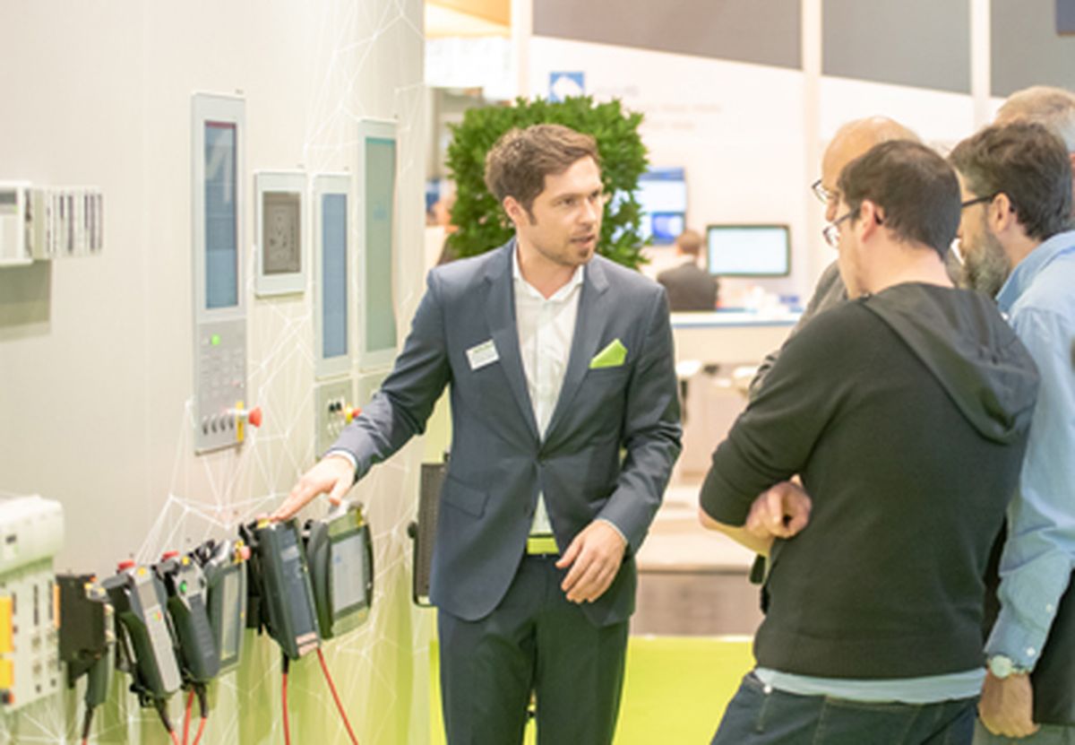 A person explaining industrial HMI and control solutions to several visitors using displayed devices at a trade fair booth.