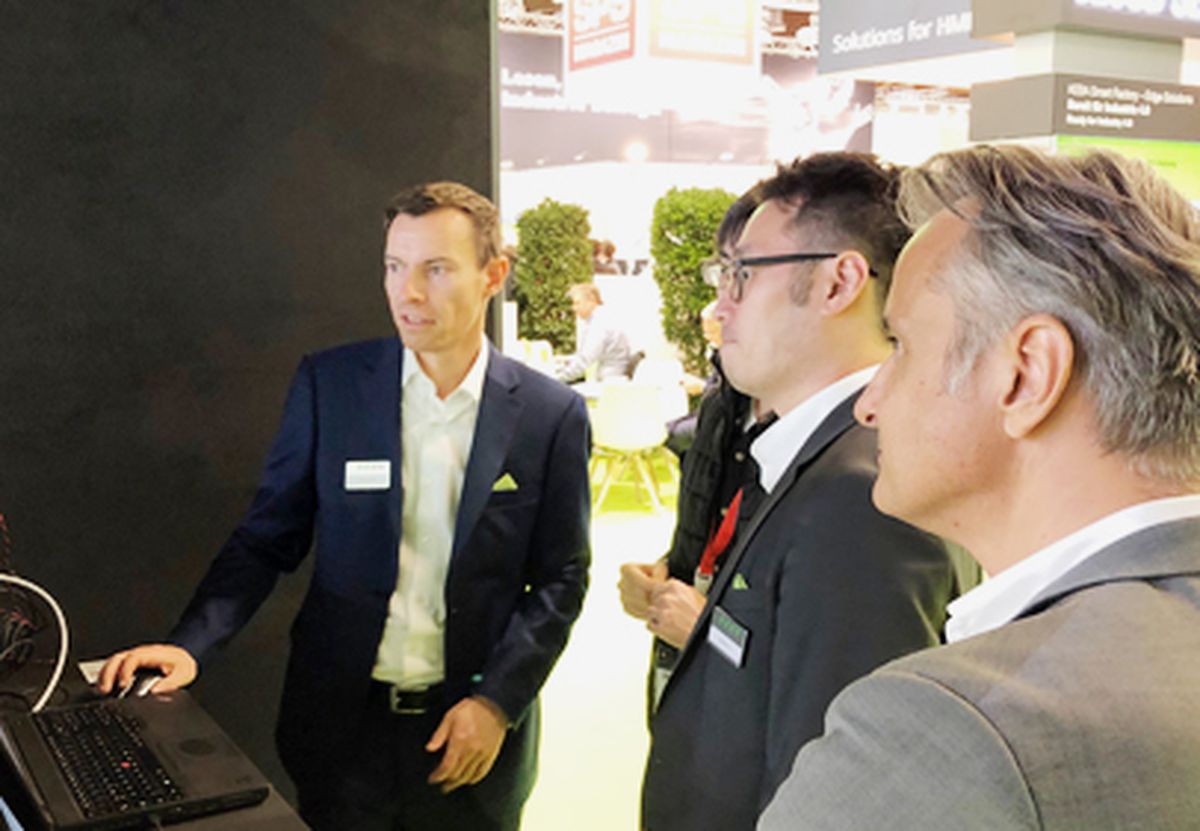 A person explaining a software-based automation or data solution to several visitors at a trade fair booth, supported by a live demonstration on a computer.