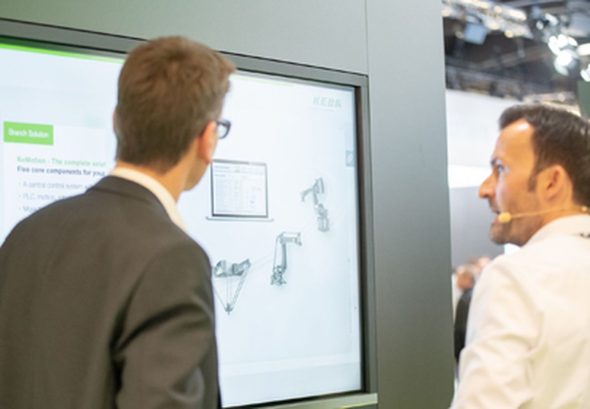 Two people viewing a presentation with graphical representations of robotics or engineering content on a large screen at a trade fair.