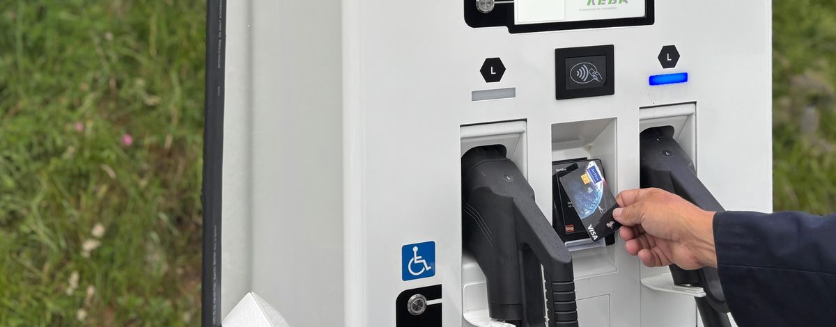 Person, die mit einer Karte an einer KEBA-Ladestation für Elektrofahrzeuge bezahlt, mit zwei sichtbaren Ladeanschlüssen. Im Hintergrund ist Gras zu sehen.