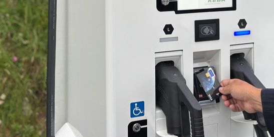 Persona che paga con una carta presso una stazione di ricarica per veicoli elettrici KEBA, con due connettori di ricarica visibili. Sullo sfondo c’è dell’erba.