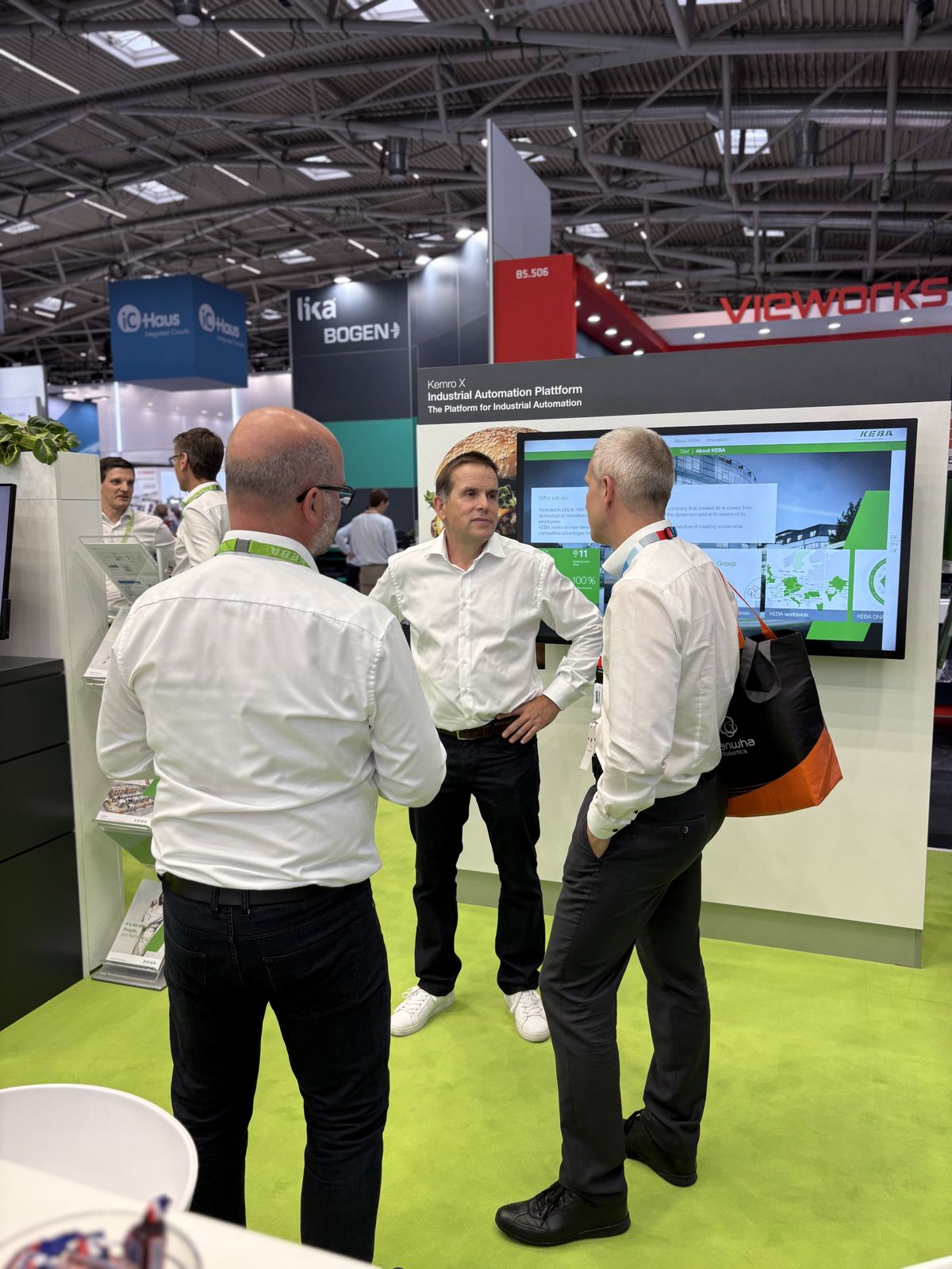 Drei Männer führen ein Gespräch am KEBA-Messestand vor einem Bildschirm mit Informationen zur Kemro X Plattform.