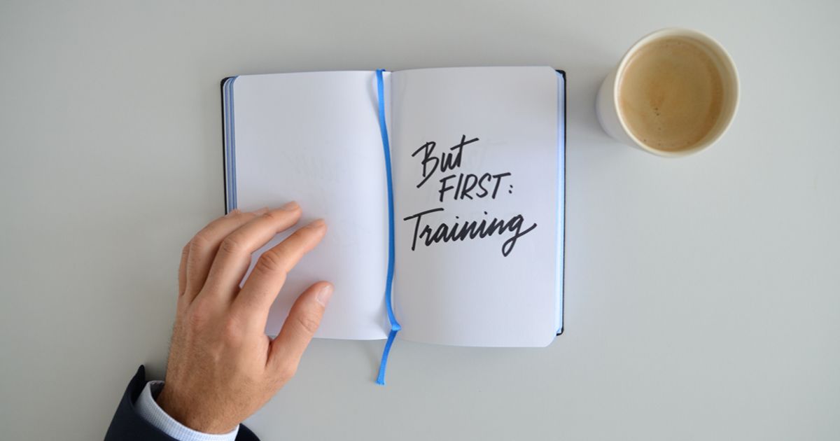 Overhead view of a CEO’s hand opening a notebook with the handwritten words “But first: Training,” with a cup of coffee beside it on a clean desk.