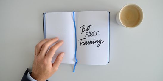 Draufsicht auf die Hand eines CEOs, der ein Notizbuch öffnet, auf dem handschriftlich „But first: Training“ steht; daneben steht eine Tasse Kaffee auf einem aufgeräumten Schreibtisch.