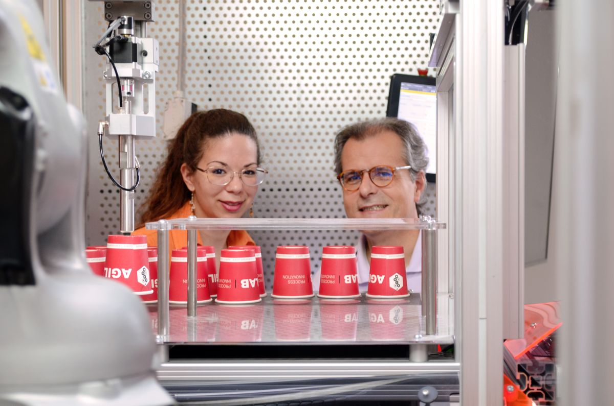 Zwei Personen beobachten eine automatisierte Roboteranwendung, bei der rote Becher mit dem KEBA-Logo sortiert werden. Das Bild zeigt die Zusammenarbeit von Mensch und Maschine in einer modernen Labor- oder Testumgebung.