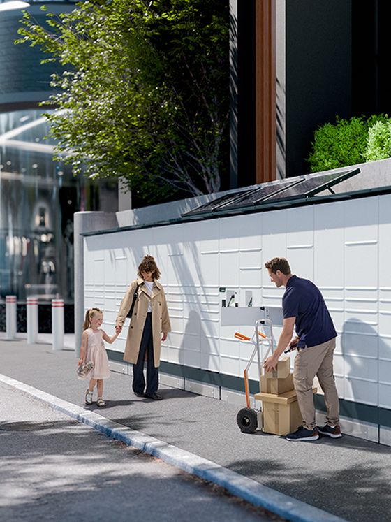A modern locker system with a solar panel on top, situated near a playground and surrounded by tall buildings under a partly cloudy sky.