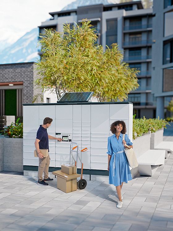 A modern parcel locker with a solar panel on top, situated in a paved courtyard with trees and buildings in the background.