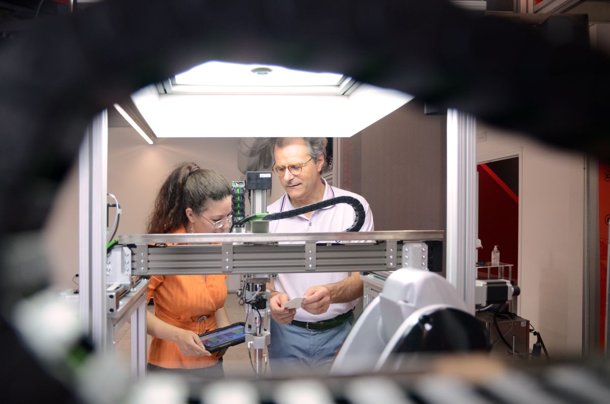 Zwei Personen arbeiten an einer automatisierten Teststation mit einem Industrieroboter. Eine Person bedient ein Tablet, während die andere eine Komponente überprüft – Darstellung präziser Zusammenarbeit zwischen Mensch und Maschine in der Automatisierung.