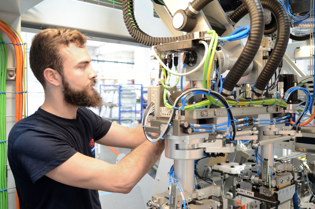 Tecnico che regola un impianto di produzione robotizzato con cablaggi, tubi pneumatici e sensori visibili. L’immagine rappresenta l’automazione di precisione e la stretta collaborazione tecnica tra SACMI e KEBA.