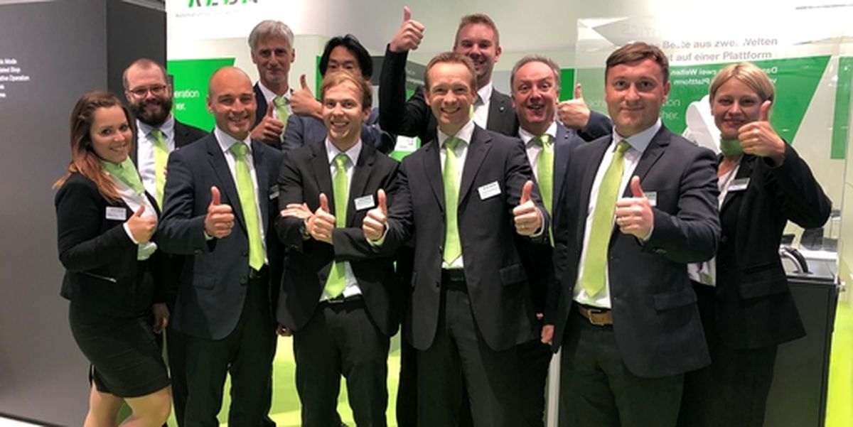 Mehrere Personen in Businesskleidung stehen dicht beieinander und zeigen mit erhobenem Daumen Zustimmung. Im Hintergrund sind KEBA-typische grüne Gestaltungselemente und Messegrafiken zu sehen. Das Bild vermittelt Teamgeist, Erfolg und positive Stimmung im Kontext eines Messeauftritts im Bereich industrieller Automatisierung.