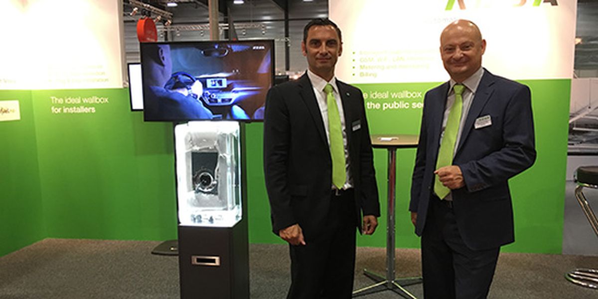 Two men in suits with green ties standing at a KEBA exhibition booth featuring a wall box display and a screen in the background.