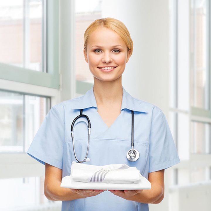 Smiling healthcare worker in blue uniform holds folded towels, standing near large windows in a bright room, with a stethoscope around her neck.
