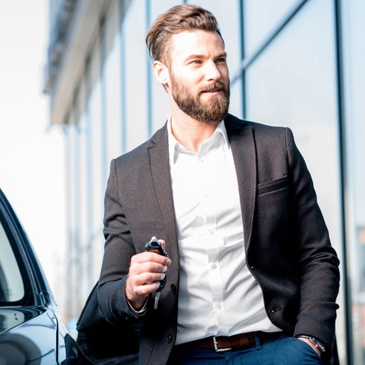 A man in a suit with a beard holds car keys, leaning against a car. A modern glass building is in the background.