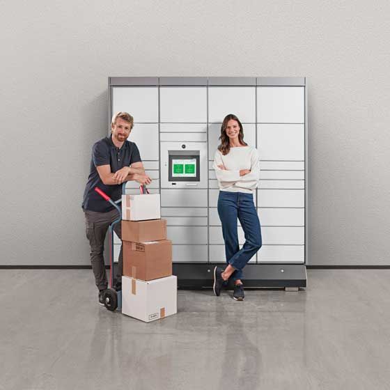 Un bărbat cu un cărucior încărcat și o femeie cu brațele încrucișate stau în fața unei stații de colete KEBA.