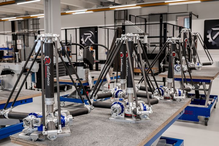 Several identical autonox robot arms mounted on work tables in a modern factory hall, with machines in the background and the company logo on the wall.