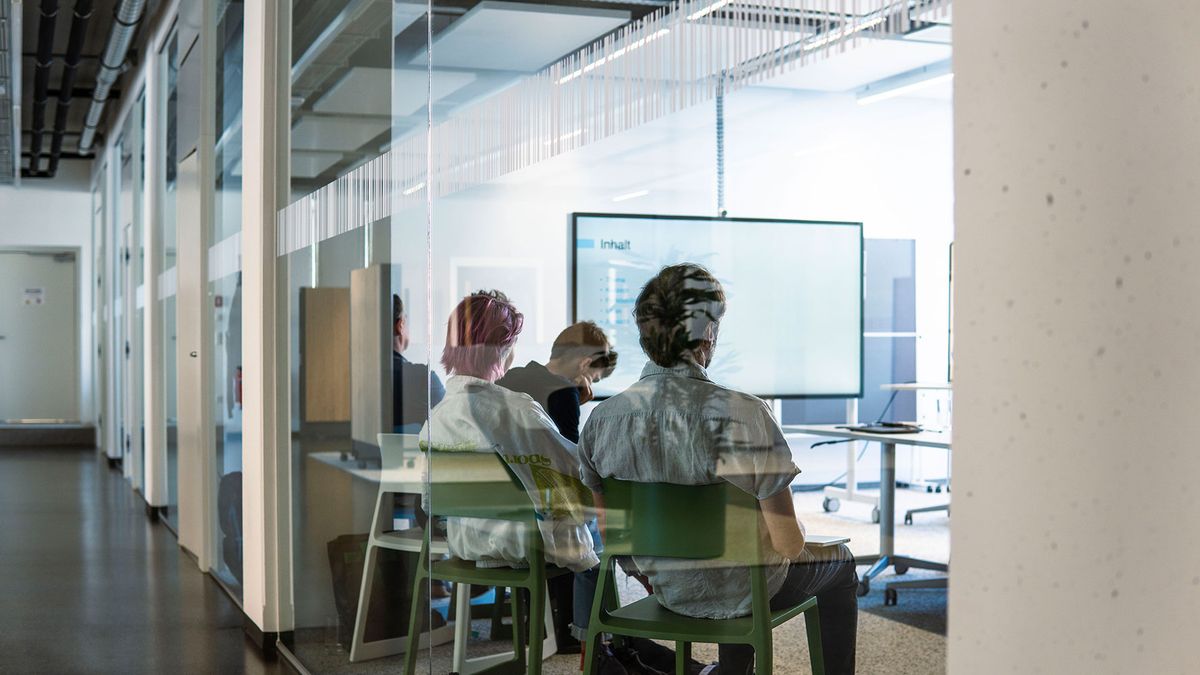 Behind a glass wall, young people are participating in a modern training session – the image conveys focused learning, digital presentation technology, and an open, collaborative learning environment.