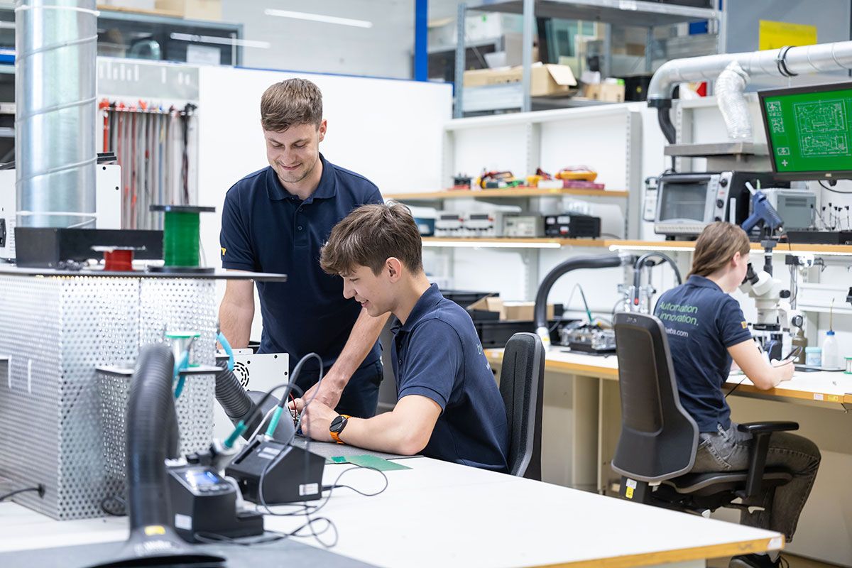 Drei Mitarbeitende arbeiten in einem Elektronik- und Automatisierungslabor an technischen Komponenten und Baugruppen. Zwei Personen prüfen gemeinsam eine Baugruppe am Arbeitsplatz, während eine weitere Person im Hintergrund an einem Mikroskop arbeitet.