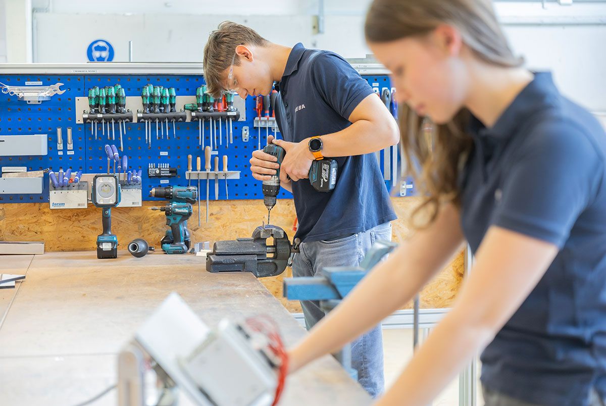 Zwei junge Mitarbeitende arbeiten in einer technischen Werkstatt an Werkstücken und Bauteilen. Im Hintergrund bohrt eine Person mit einem Akkuschrauber ein eingespanntes Metallteil, während Werkzeug und Arbeitsmittel übersichtlich an der Werkbank angeordnet sind.