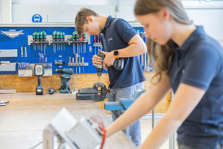 Zwei junge Mitarbeitende arbeiten in einer technischen Werkstatt an Werkstücken und Bauteilen. Im Hintergrund bohrt eine Person mit einem Akkuschrauber ein eingespanntes Metallteil, während Werkzeug und Arbeitsmittel übersichtlich an der Werkbank angeordnet sind.