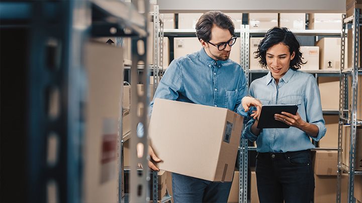 Two employees in a warehouse check delivery data on a tablet, one is holding a large box.