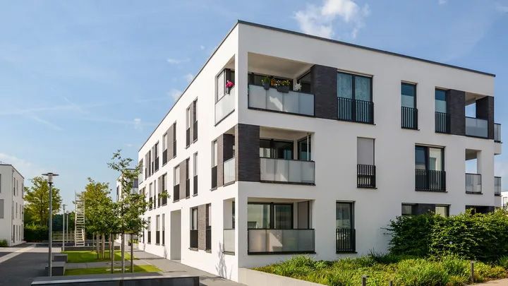Modern white apartment building with balconies and greenery on a sunny day.