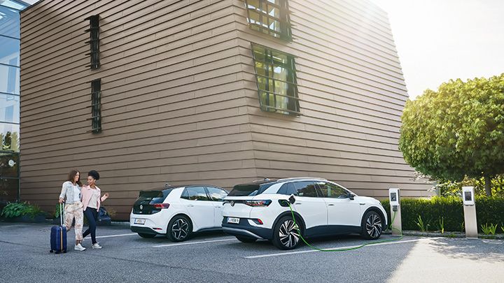 Two electric cars are charging at stations in front of a modern building while two women walk by with suitcases.
