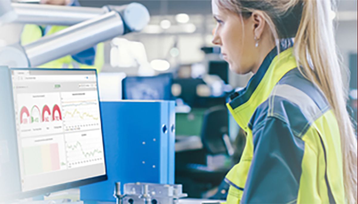 Female technician wearing a safety vest working at an industrial workstation, analyzing production and process data on a monitor. The display shows charts and key performance indicators for quality monitoring and automated manufacturing performance.