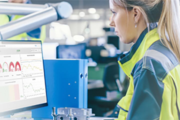 Female technician wearing a safety vest working at an industrial workstation, analyzing production and process data on a monitor. The display shows charts and key performance indicators for quality monitoring and automated manufacturing performance.
