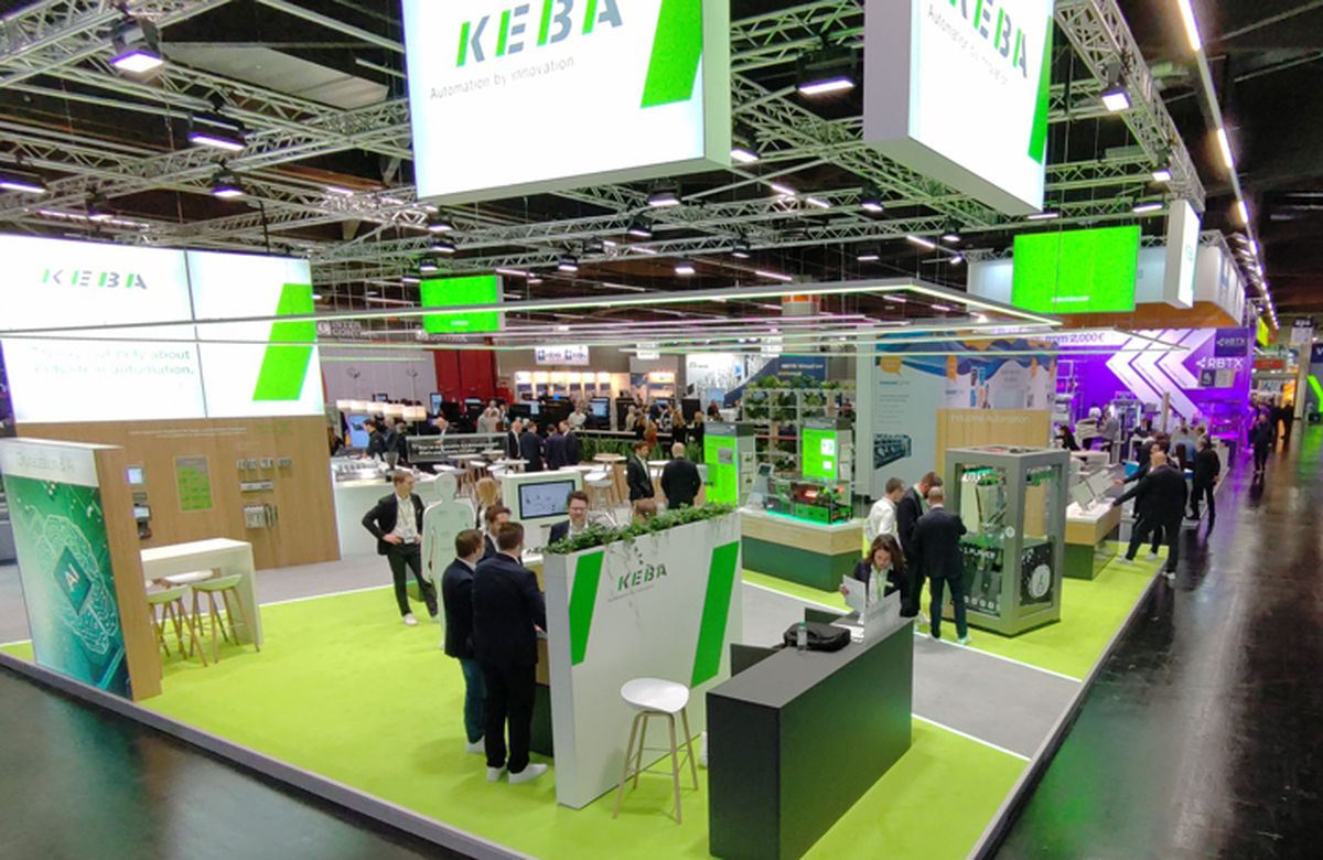 Wide-angle view of a large trade fair booth with presentation areas, product demonstrations, and visitors in an industrial automation context.