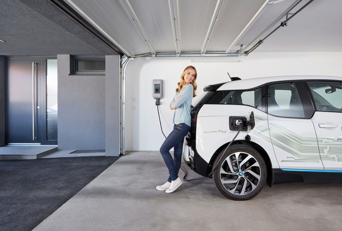 Woman leaning casually against an electric car in a garage while it is charging at a wallbox.