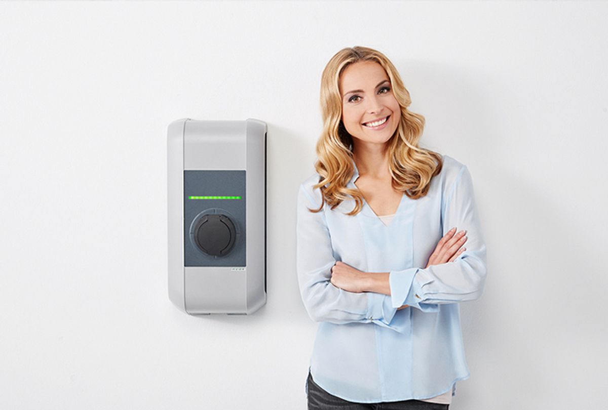 Smiling woman standing next to a mounted wallbox on a white wall.