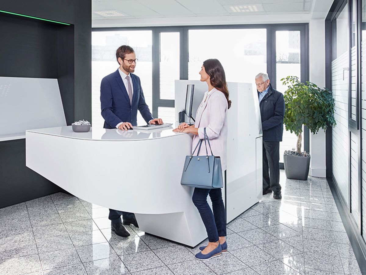 In einer modernen Bankfiliale spricht eine Frau mit einem Mitarbeiter am Empfang, während ein Mann im Hintergrund einen Bankautomaten bedient.