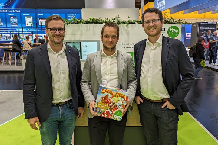 Three people standing together at a trade fair booth holding a prize or promotional item as part of an exhibition activity.