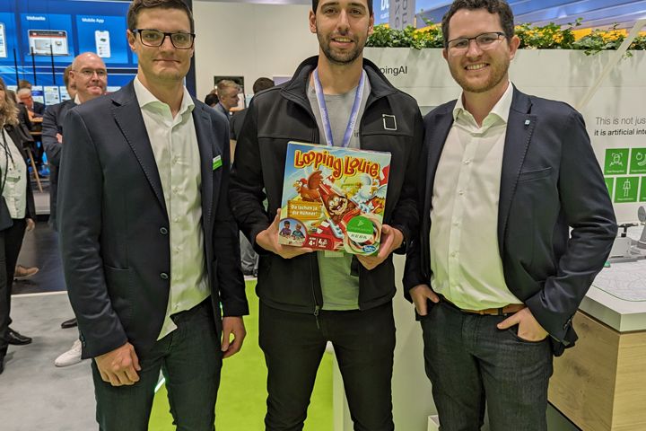 Three people standing together at a trade fair booth holding a prize or promotional item as part of an exhibition activity.