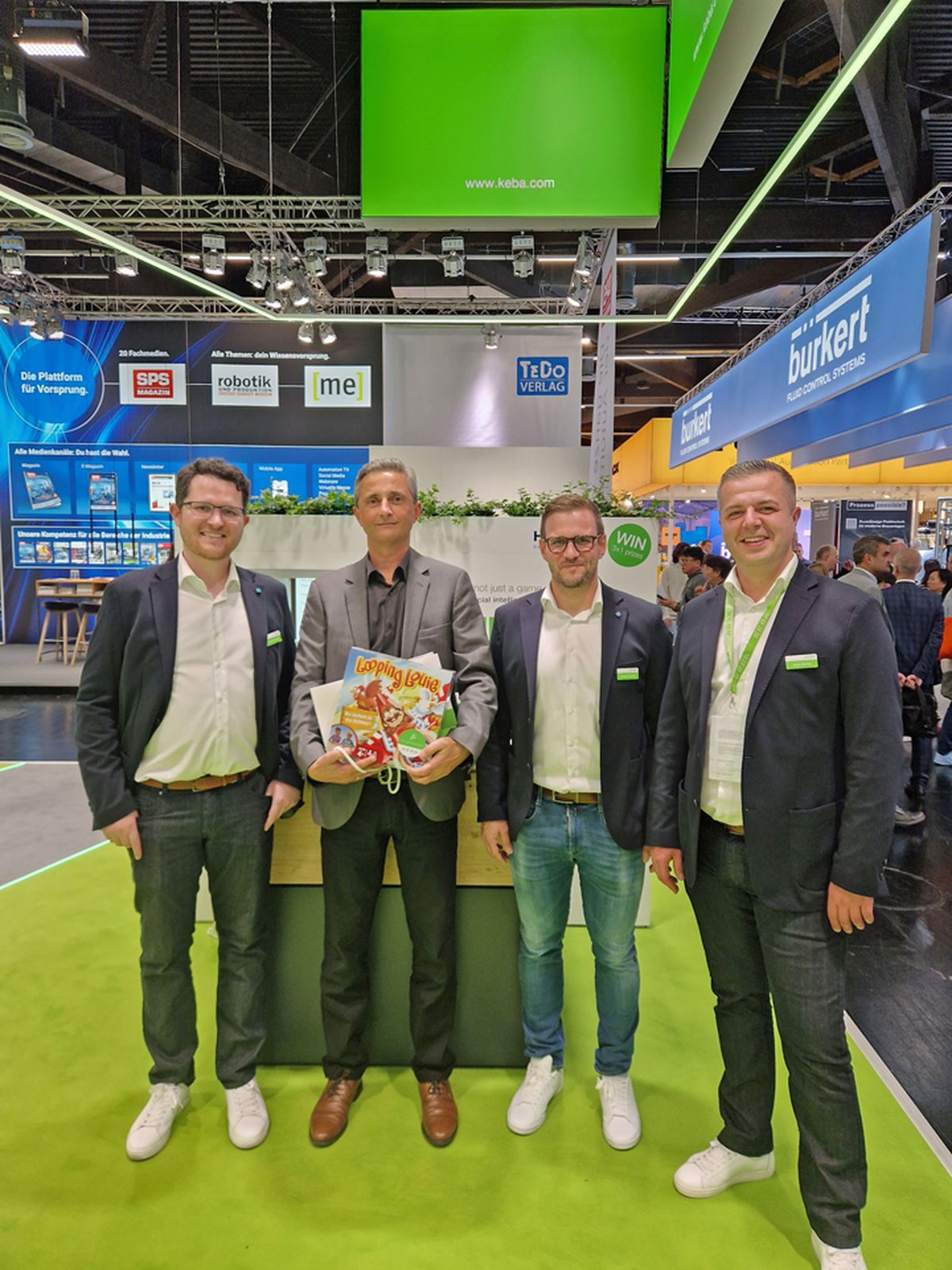 Three people standing together at a trade fair booth holding a prize or promotional item as part of an exhibition activity.