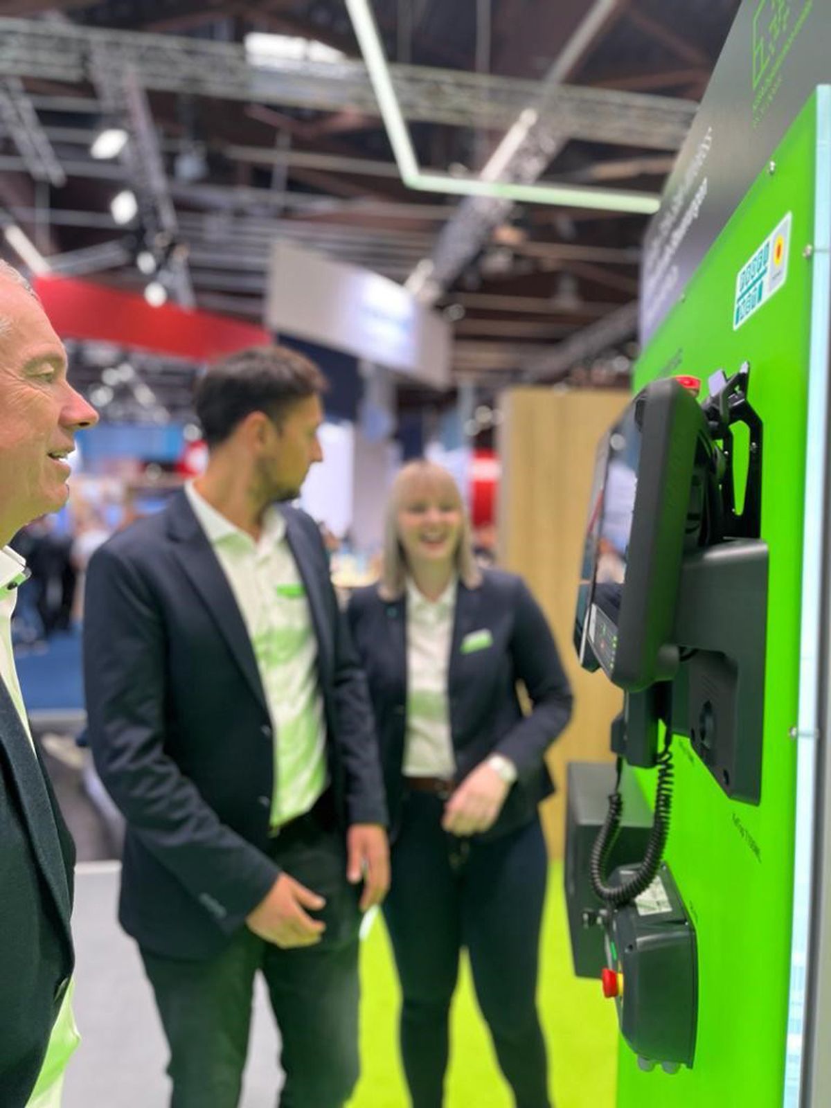Several people examining and discussing a mounted HMI operator device during a product demonstration at a trade fair booth.