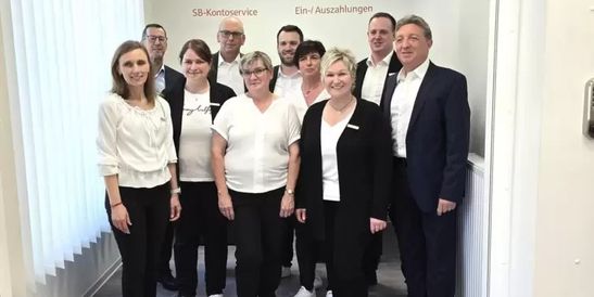 Gruppe von zehn Personen in einem Raum, teils formell, teils leger gekleidet, lächeln in die Kamera. Im Hintergrund ist der KI-Assistent KeBob zu sehen.