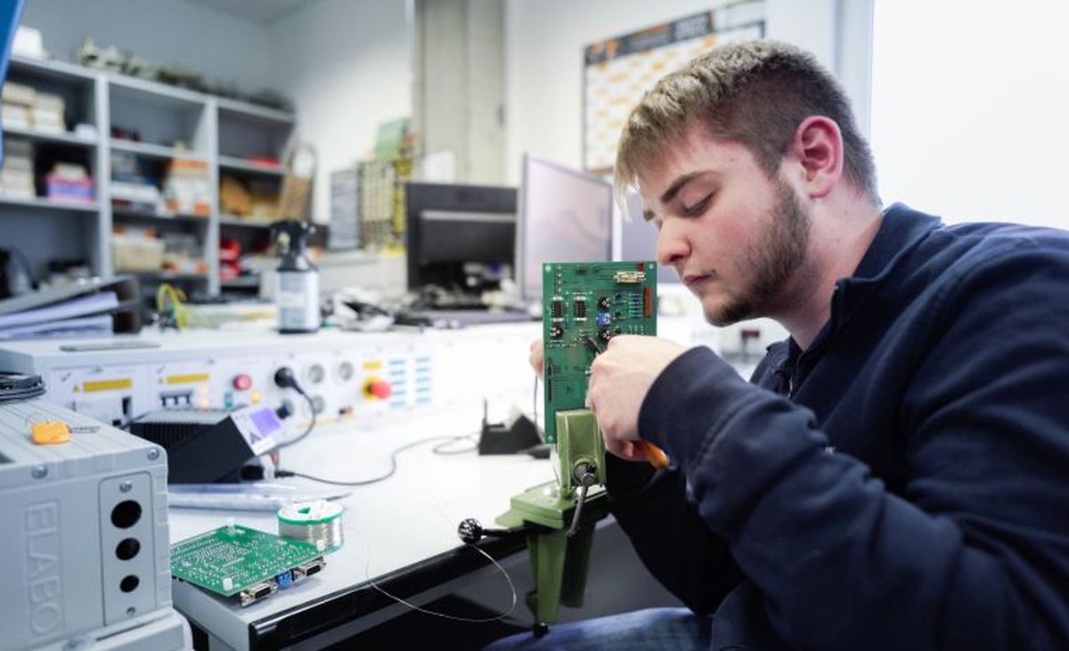 Ein KEBA-Auszubildender lötet präzise an einer senkrecht eingespannten Leiterplatte in einem Elektroniklabor mit professioneller Arbeitsplatzausstattung.