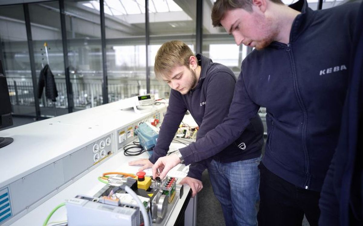 Zwei KEBA-Auszubildende arbeiten gemeinsam an einem elektrischen Versuchsaufbau mit Steuerungstechnik und Not-Aus-Schalter in einem modern ausgestatteten Labor.