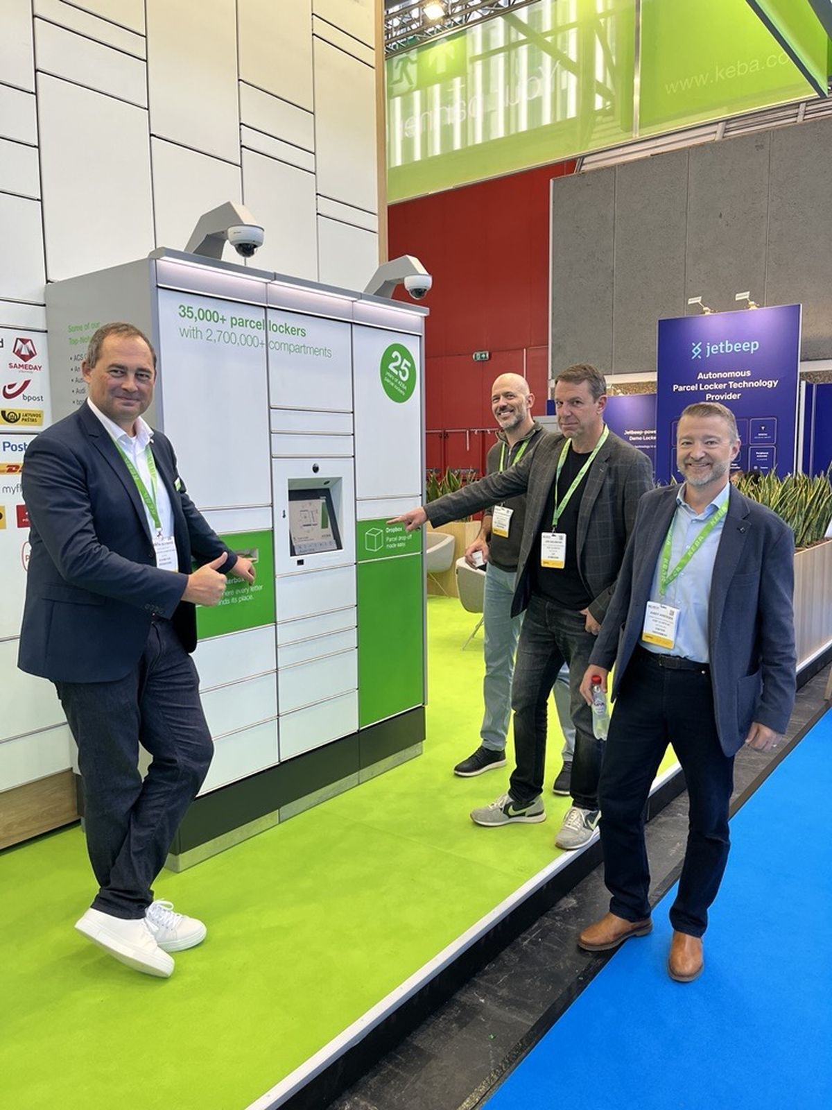 Four men stand near a green and white parcel locker display at a Parcel + Post Expo 2025, smiling and gesturing towards it.