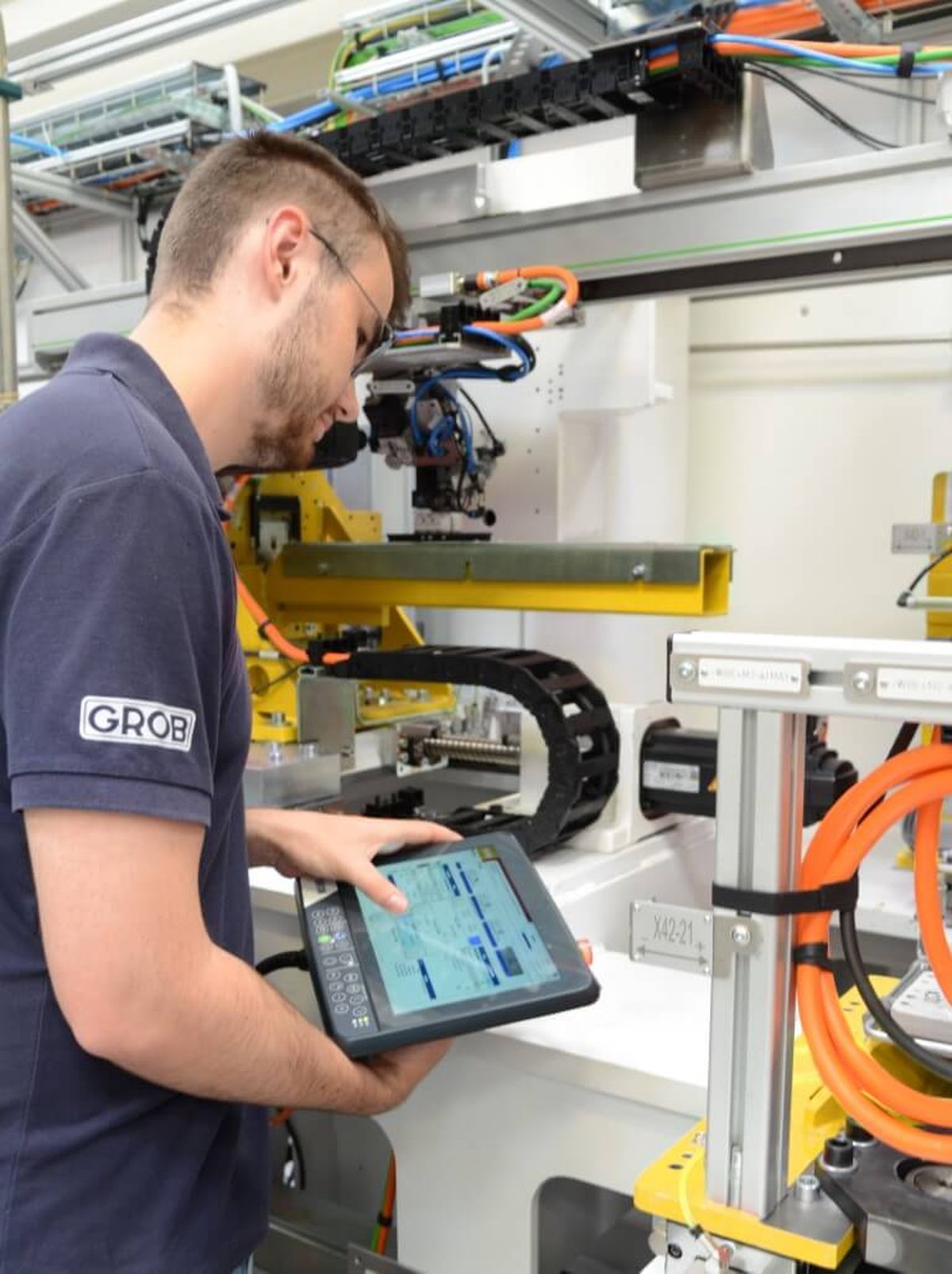 A technician operates an automated production system by GROB using a mobile control panel. The image represents intelligent machine control, digitalization, and efficient process automation.