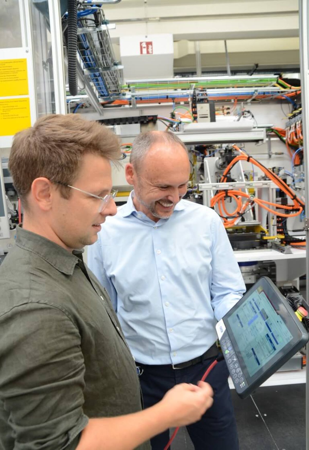 Two men standing in front of an automated system operating a mobile touchscreen control device. The image symbolizes teamwork, expertise, and digital machine control in industrial automation.
