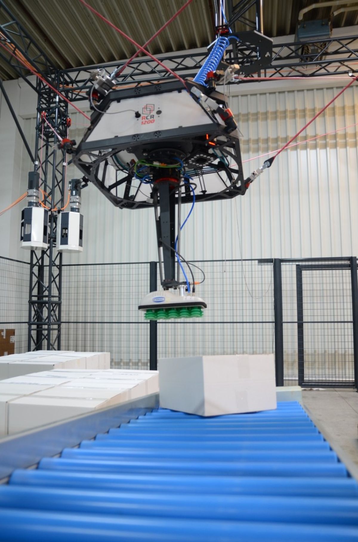 A cable robot picks up a box from an inclined roller conveyor with its platform tilted in a controlled manner. The adjustable tilt allows precise handling without the need for an additional axis as required by gantry systems.