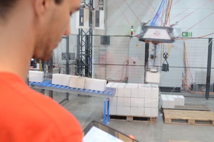 An operator controls a cable robot in an industrial hall using a wireless handheld device with a large display. The image illustrates ergonomic and safe robot programming via wireless HMI while maintaining a clear view of the system.